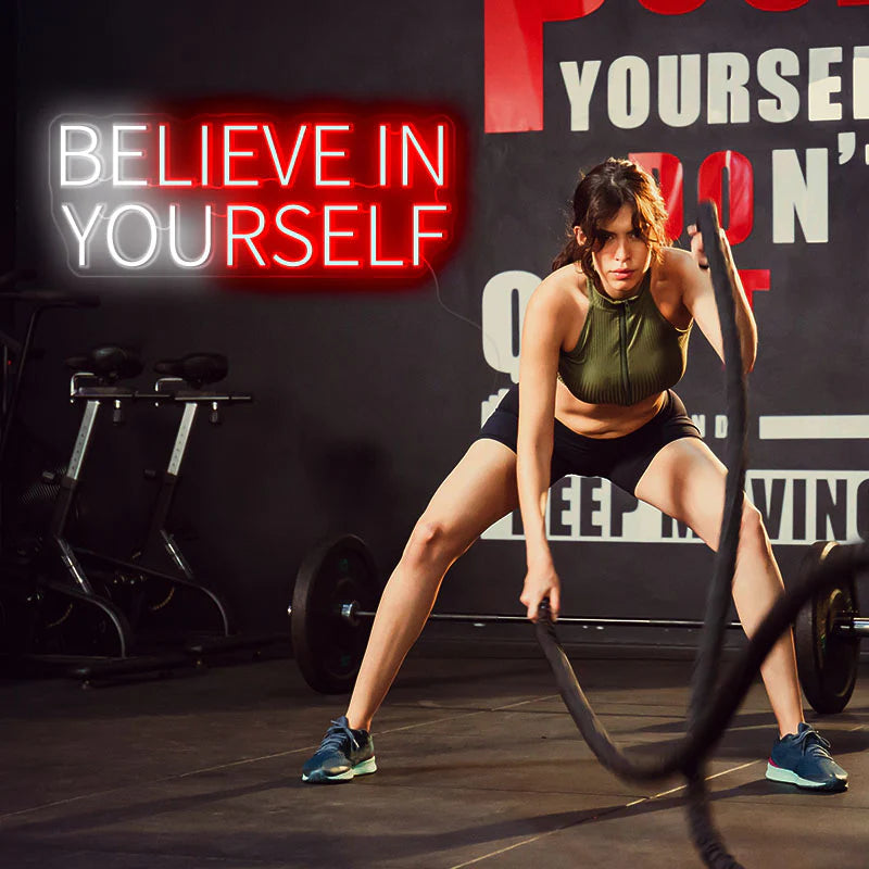BELIEVE IN YOURSELF neon sign. Sign is in red and white. On the sign's right, there is a female working out with ropes. The location of the image is in a gym. 