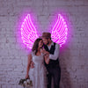 Wedding Couple posing infront of a deep Pink Ascended Angel Wings LED neon sign which is installed on a white brick wall.