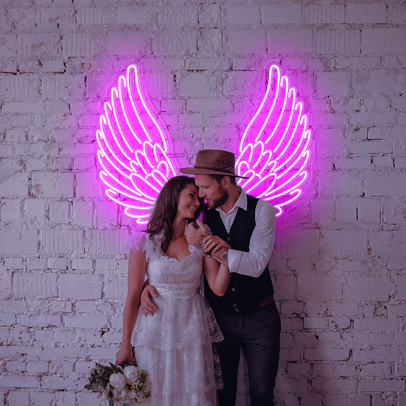 Wedding Couple posing infront of a deep Pink Ascended Angel Wings LED neon sign which is installed on a white brick wall. 